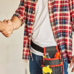 Uścisk dłoni dwóch mężczyzn w koszulach w kratę, jeden z pasem narzędziowym, na tle surowego wnętrza w trakcie remontu.