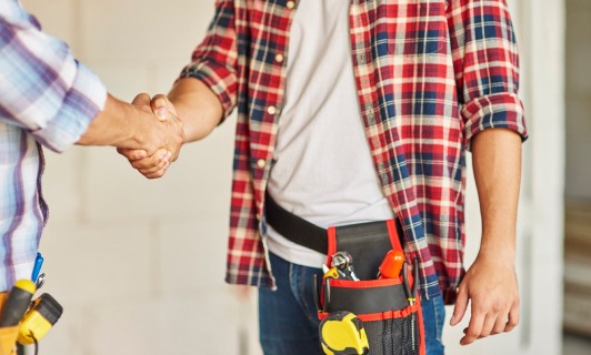 Uścisk dłoni dwóch mężczyzn w koszulach w kratę, jeden z pasem narzędziowym, na tle surowego wnętrza w trakcie remontu.