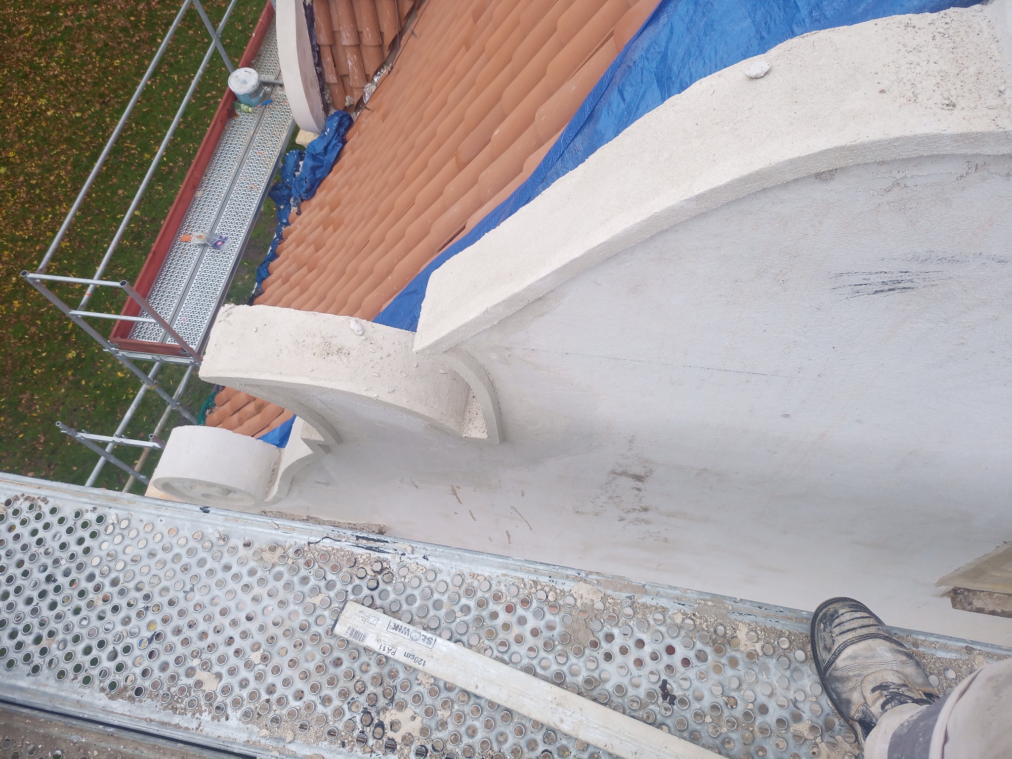 Fragment elewacji budynku w trakcie prac wykończeniowych, widoczne detale architektoniczne, rusztowanie, zabezpieczona dachówka i but robotnika.