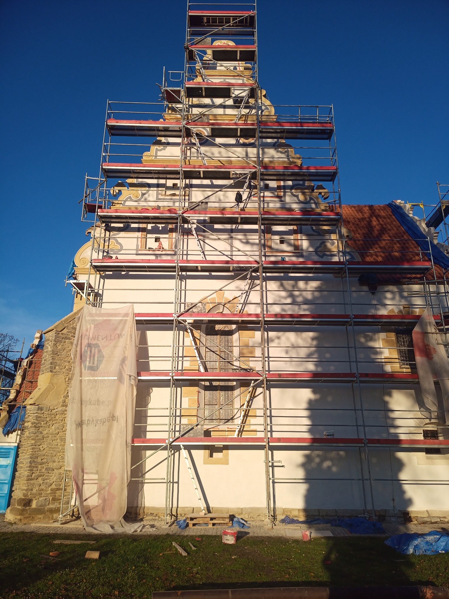 Wysoka wieża kościelna w rusztowaniach, z widocznymi elementami dekoracyjnymi i fragmentami odnowionej elewacji, częściowo zakryta siatką ochronną.