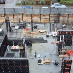 Widok z g&oacute;ry na plac budowy z przygotowanymi szalunkami fundament&oacute;w, zbrojeniami i deskowaniem ziemnym, częściowo wypełniony błotem po deszczu.