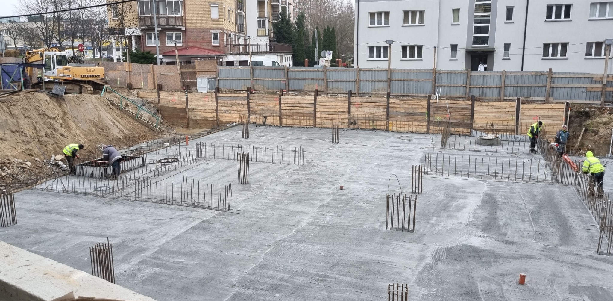 Wylany betonowy fundament z wystającymi prętami zbrojeniowymi, pracownicy w odblaskowych kamizelkach przy deskowaniu, w tle wykop i maszyna budowlana.
