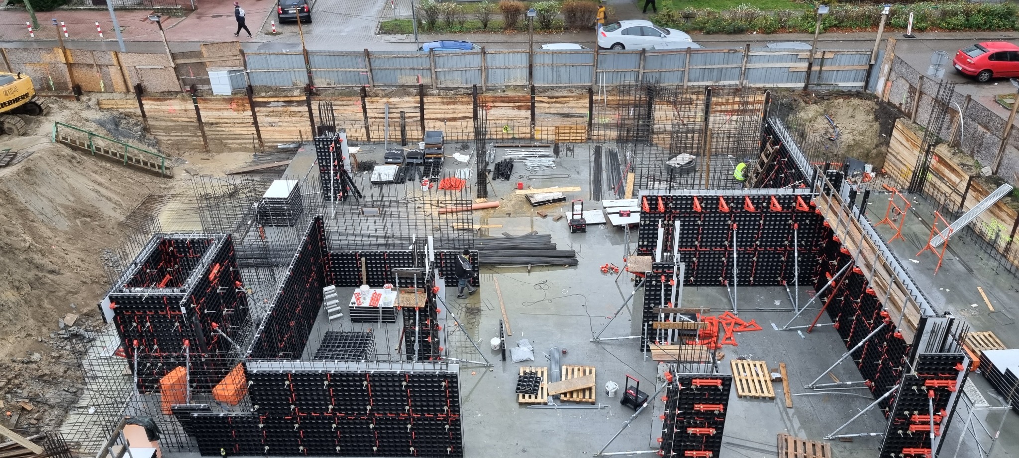 Widok z góry na plac budowy z przygotowanymi szalunkami fundamentów, zbrojeniami i deskowaniem ziemnym, częściowo wypełniony błotem po deszczu.