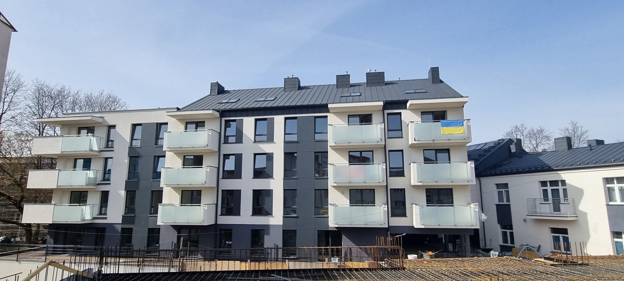 Nowoczesny budynek wielorodzinny w trakcie budowy, z widoczną konstrukcją szkieletową na pierwszym planie i powiewającą ukraińską flagą na jednym z balkonów.