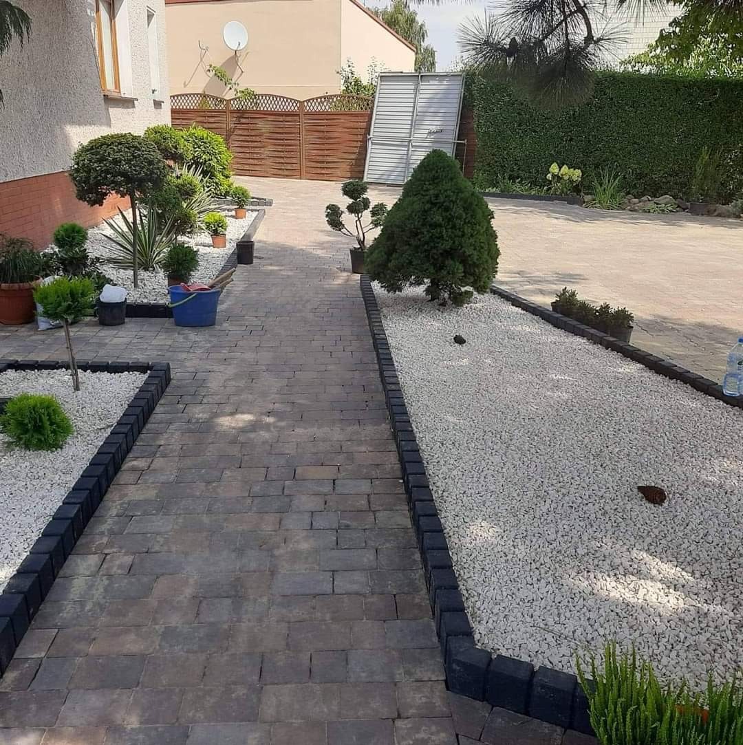 Ogród z ścieżką z kostki brukowej w odcieniach szarości, obrzeża z ciemnej kostki, rabaty z białym grysem i ozdobnymi krzewami, w tle dom z beżową elewacją i drewnianym ogrodzeniem.