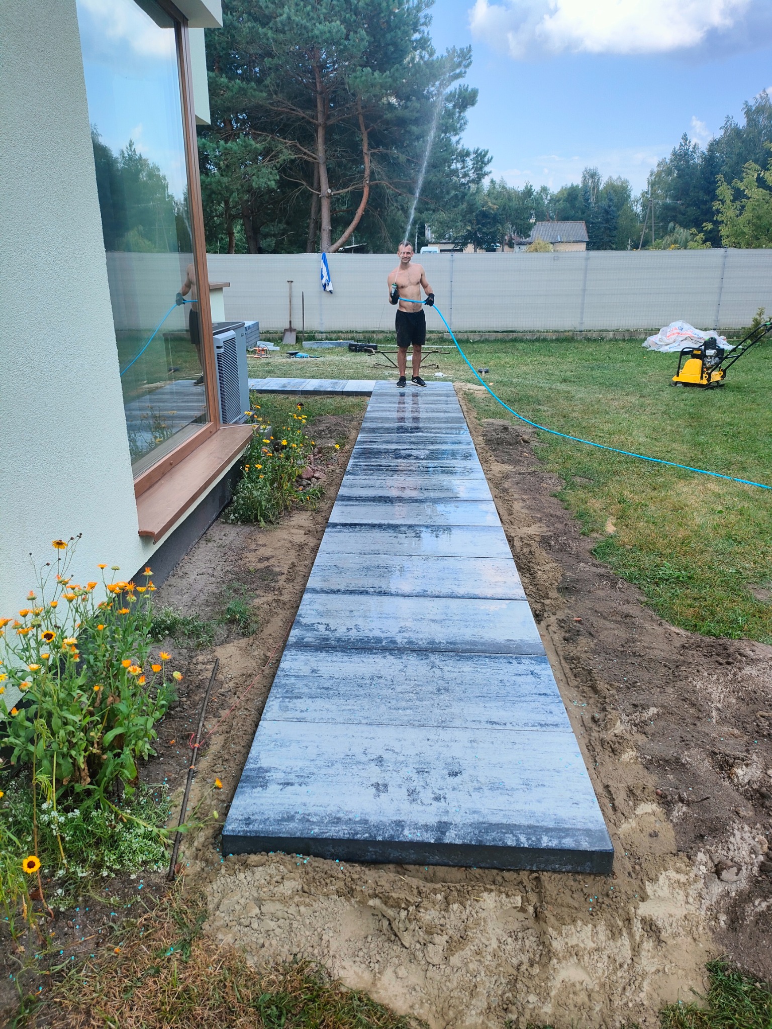 Układanie ścieżki z płyt betonowych w ogrodzie, mężczyzna podlewa wodą, widoczna zagęszczarka w tle.