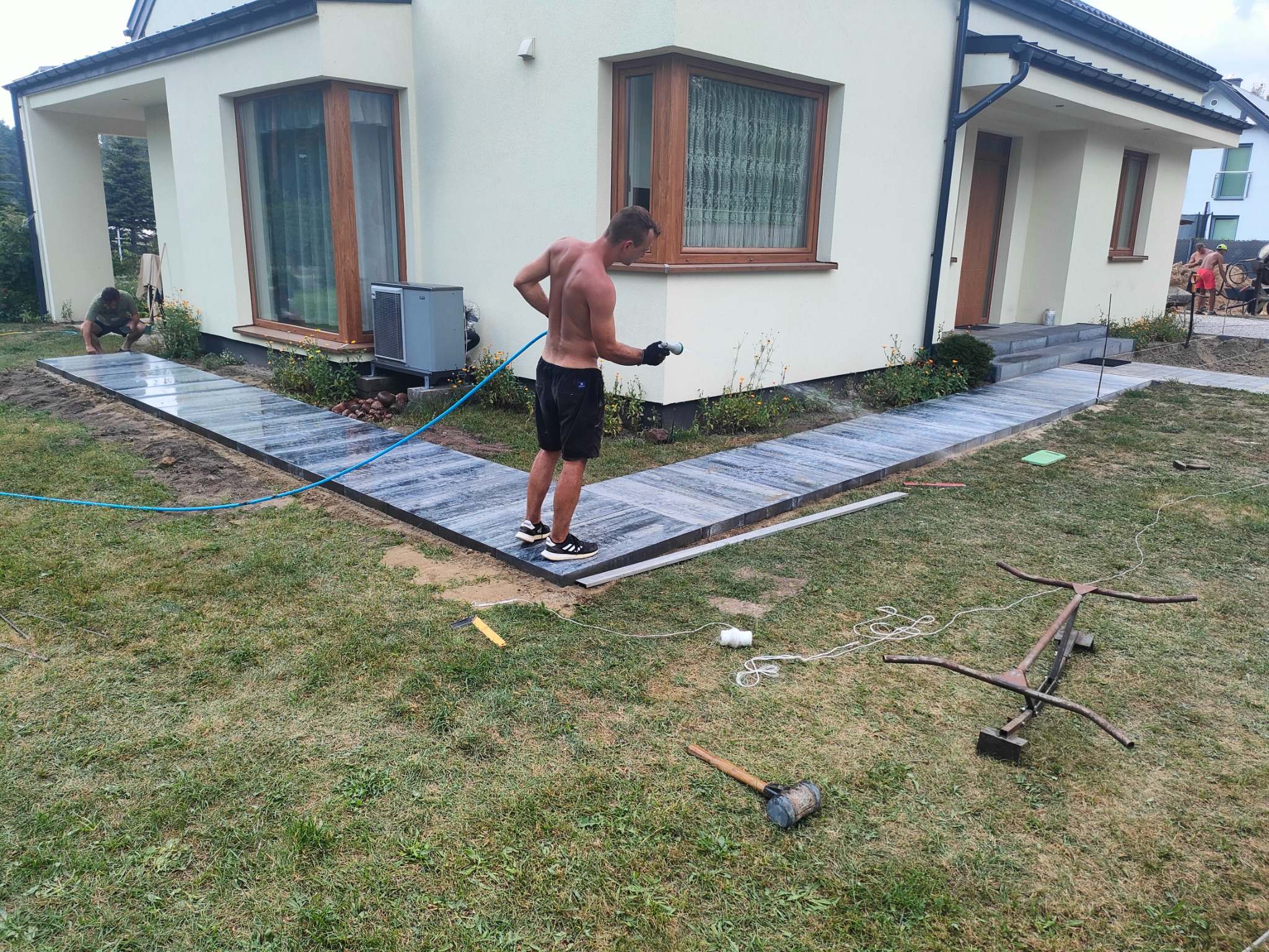 Pracownik w czarnych spodenkach i sportowych butach spryskuje wodą nowo ułożony chodnik z szarych płyt kamiennych prowadzący do domu z jasną elewacją, w tle widoczny drugi pracownik przy betoniarce.