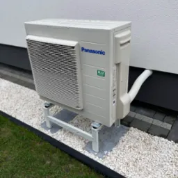 Zewnętrzna jednostka klimatyzacyjna Panasonic na metalowym stelażu, umieszczona na białym żwirze przy ścianie budynku z widoczną rurą odprowadzającą.