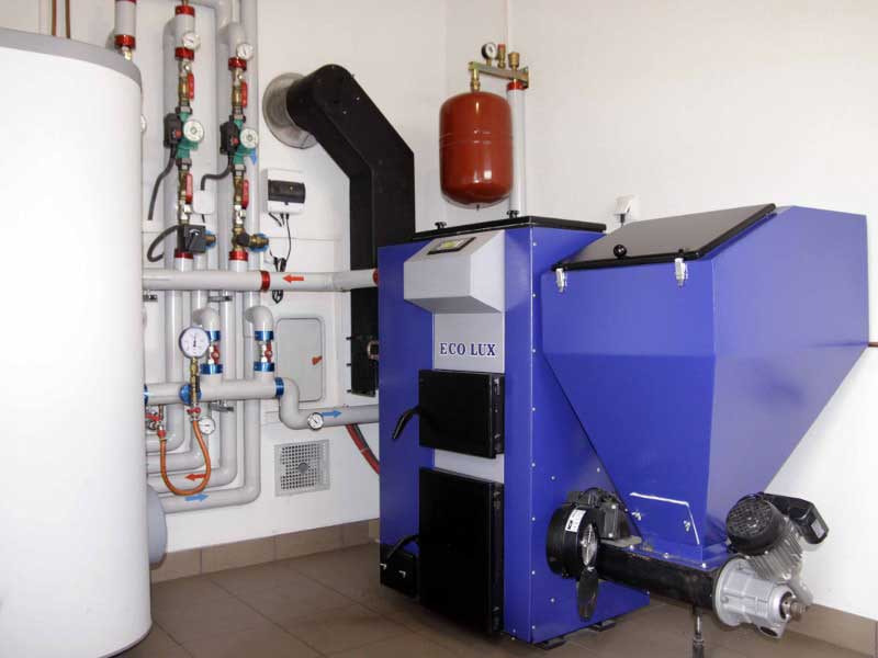 Niebieski piec na paliwo stałe Eco Lux z zasobnikiem, podłączony do instalacji CO z licznymi rurami, zaworami i manometrami, w kotłowni z białymi ścianami i brązowymi płytkami podłogowymi.