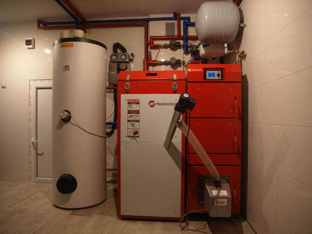 Nowoczesny piec na paliwo stałe Heitztechnik, z podajnikiem, zbiornikiem wyrównawczym i zasobnikiem ciepłej wody użytkowej w kotłowni wyłożonej białymi płytkami.