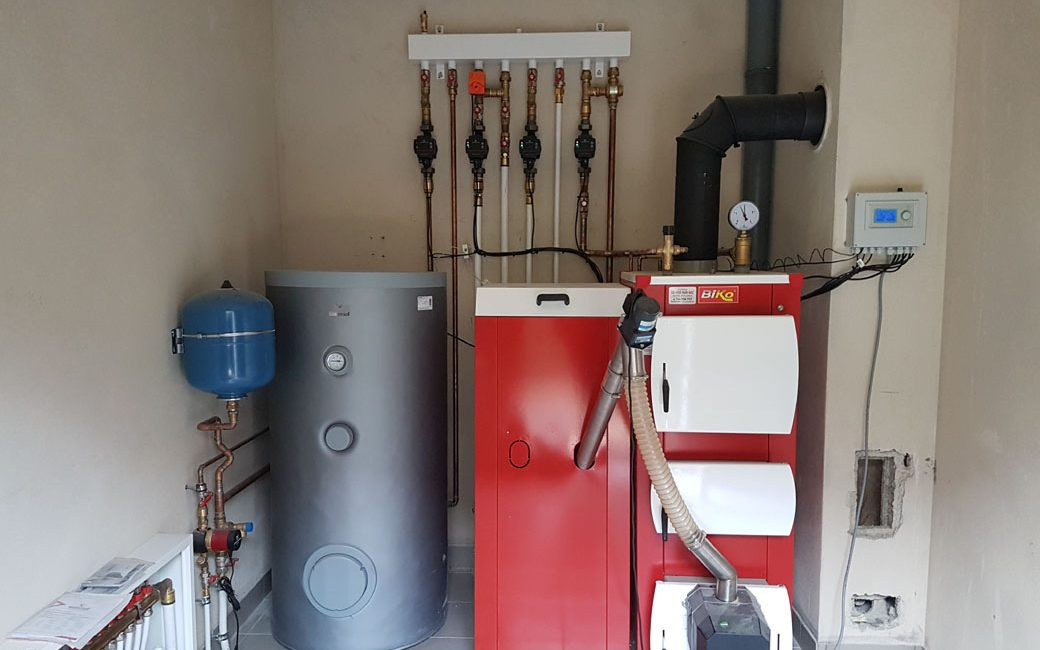 Zainstalowany system grzewczy z czerwonym piecem, szarym zbiornikiem akumulacyjnym i niebieskim naczyniem wzbiorczym w kotłowni o jasnych ścianach.