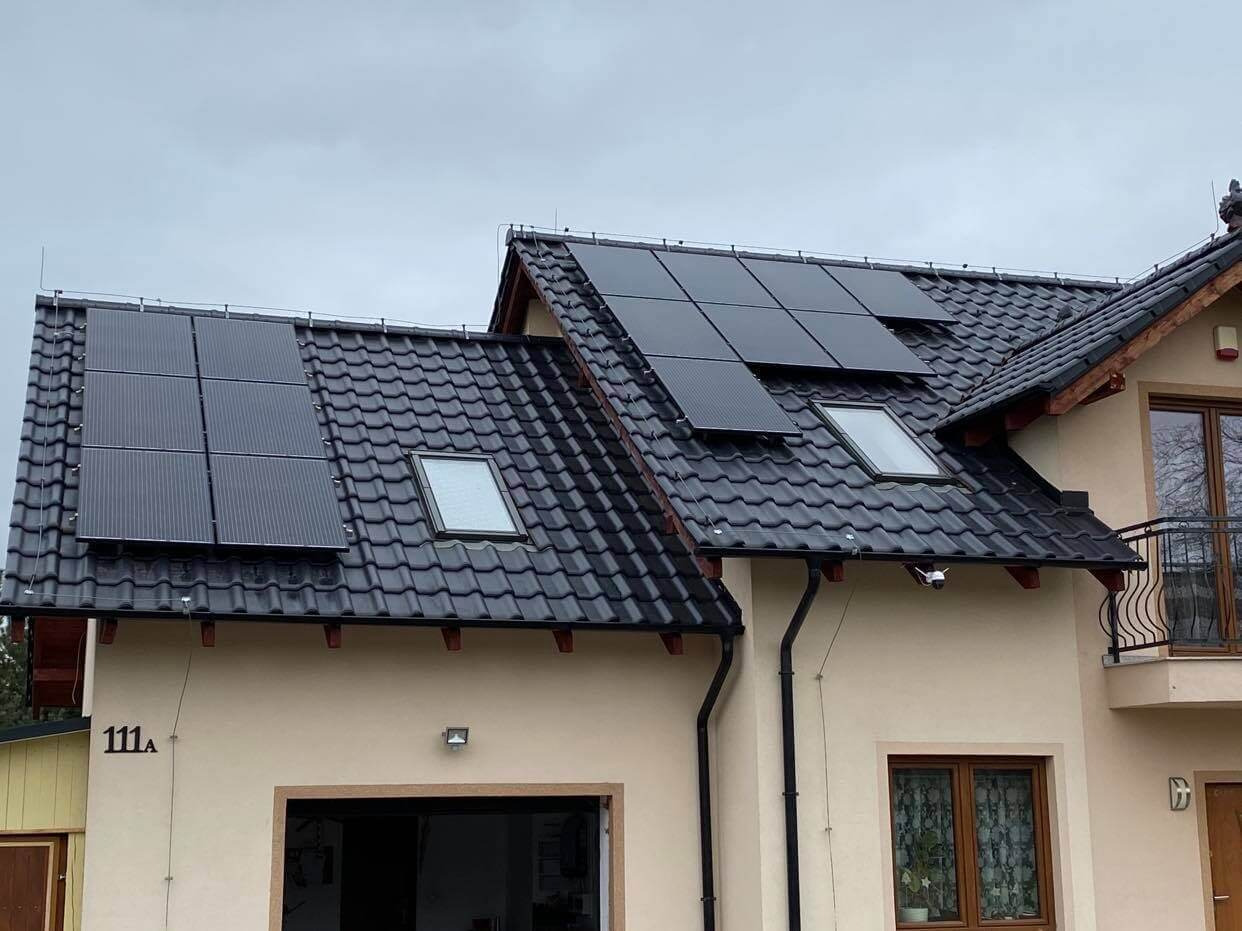 Cztery panele fotowoltaiczne na dachu domu z czarną dachówką, widoczne okna dachowe i numer domu 111A.