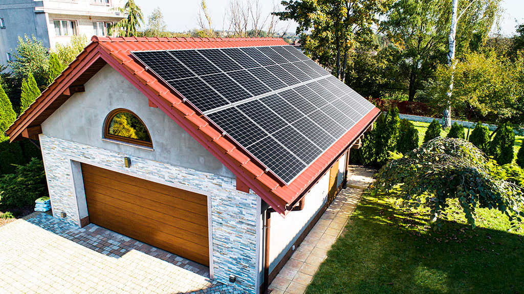 Widok z góry na garaż z panelami słonecznymi na czerwonym dachu, otoczony zielonym trawnikiem i drzewami.