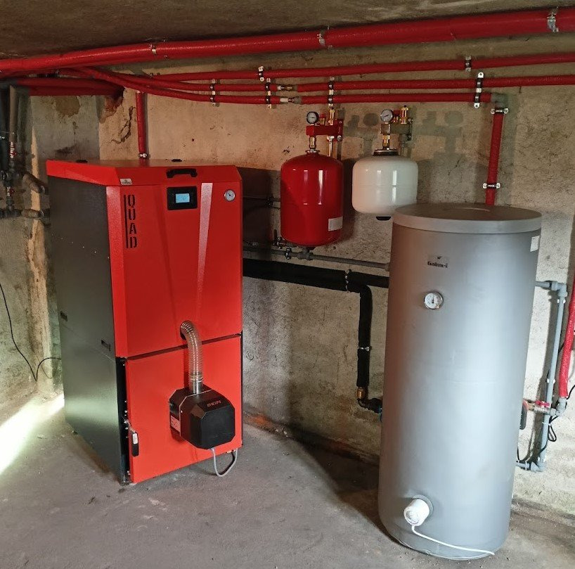 Czerwony piec na paliwo stałe marki QUAD z podajnikiem, obok szary zasobnik ciepłej wody użytkowej, czerwone rury instalacji CO zamocowane na ścianie w piwnicy.