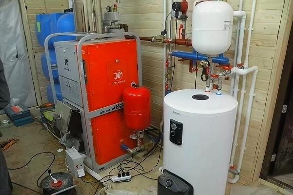 Pomarańczowo-szary piec grzewczy marki Kiturami z czerwonym zbiornikiem wyrównawczym, obok biały zasobnik ciepłej wody z podłączeniami hydraulicznymi w pomieszczeniu wyłożonym boazerią.