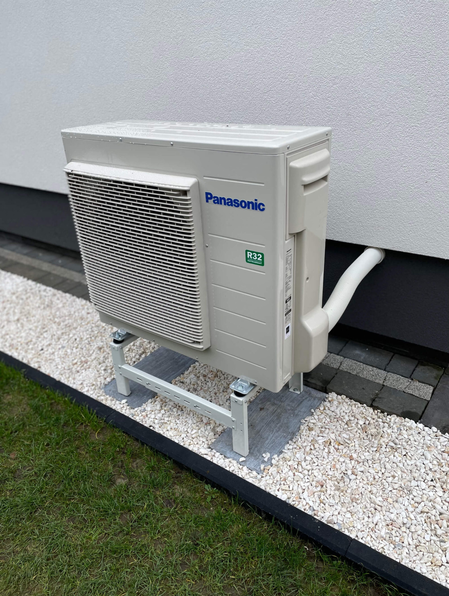Zewnętrzna jednostka klimatyzacyjna Panasonic na metalowym stelażu, umieszczona na białym żwirze przy ścianie budynku z widoczną rurą odprowadzającą.