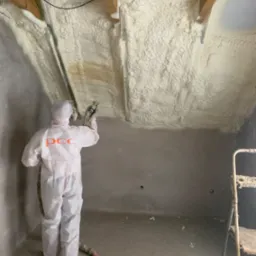 Pracownik w kombinezonie ochronnym z napisem 'PCC' aplikuje piankę izolacyjną na poddasze, widoczne drewniane elementy konstrukcji dachu i drabina.