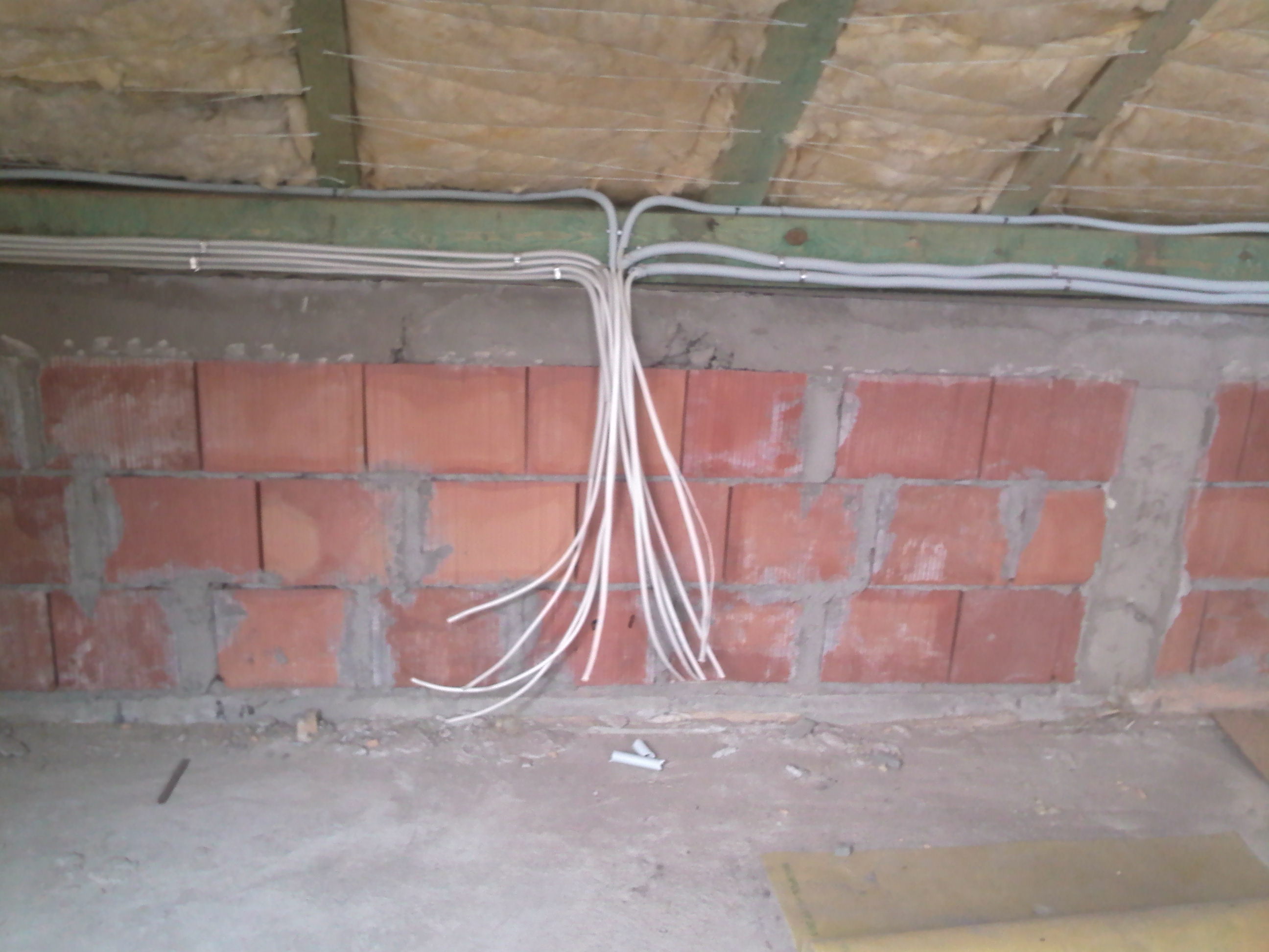 Białe przewody elektryczne wychodzące z szarej rury zamocowanej do sufitu, zwisające wzdłuż ceglanej ściany w surowym stanie.