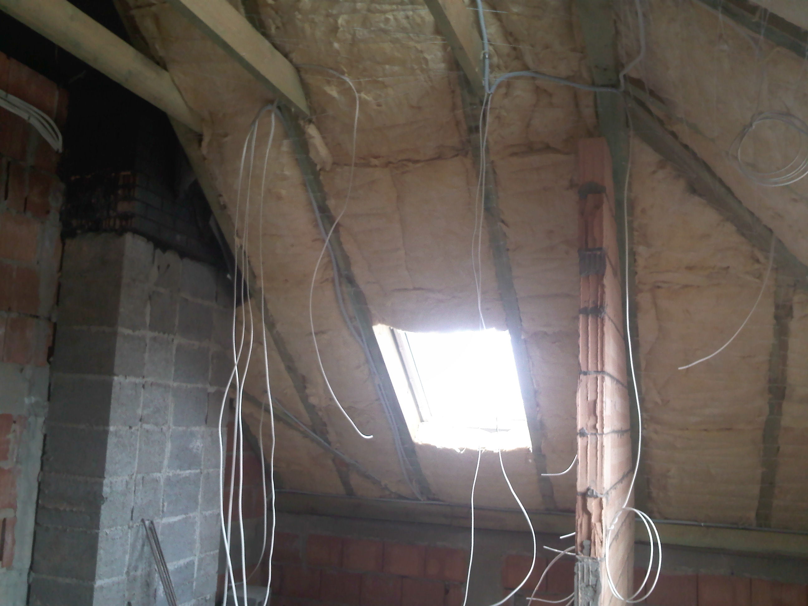 Poddasze w trakcie adaptacji: widoczne ocieplenie wełną mineralną, drewniane belki konstrukcyjne, mur z bloczków i przewody elektryczne zwisające z sufitu w kierunku okna dachowego.