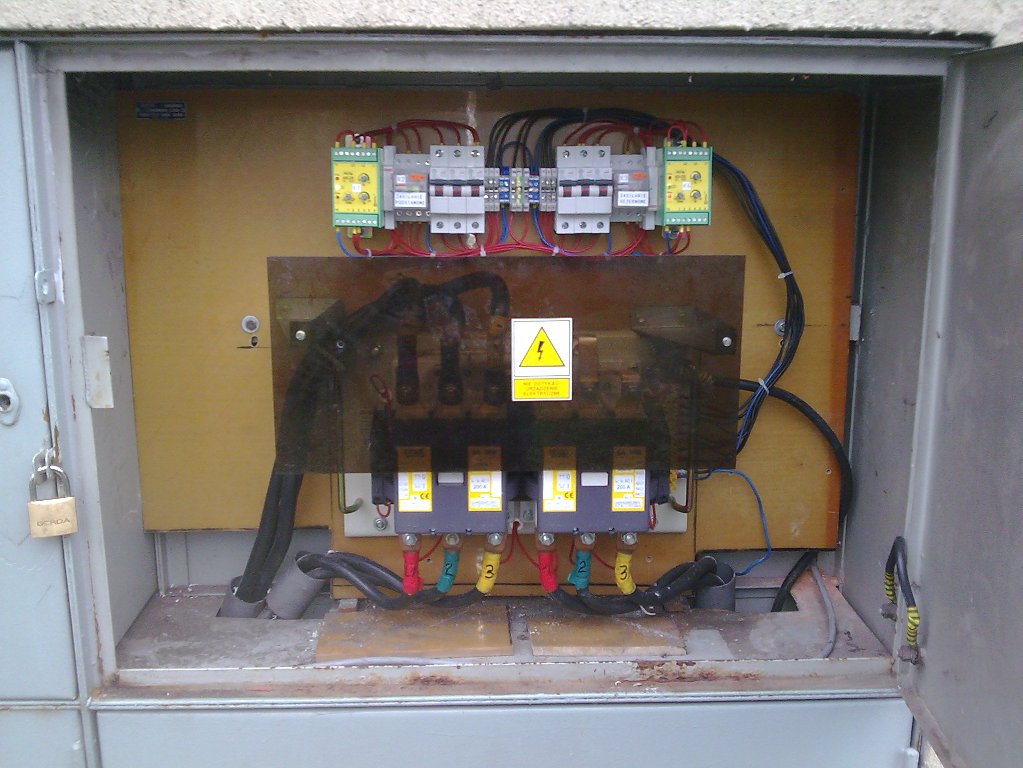 Otwarta skrzynka elektryczna z zainstalowanymi bezpiecznikami, przewodami i tabliczką ostrzegawczą, widoczne kolorowe oznaczenia przewodów i kłódka zabezpieczająca.