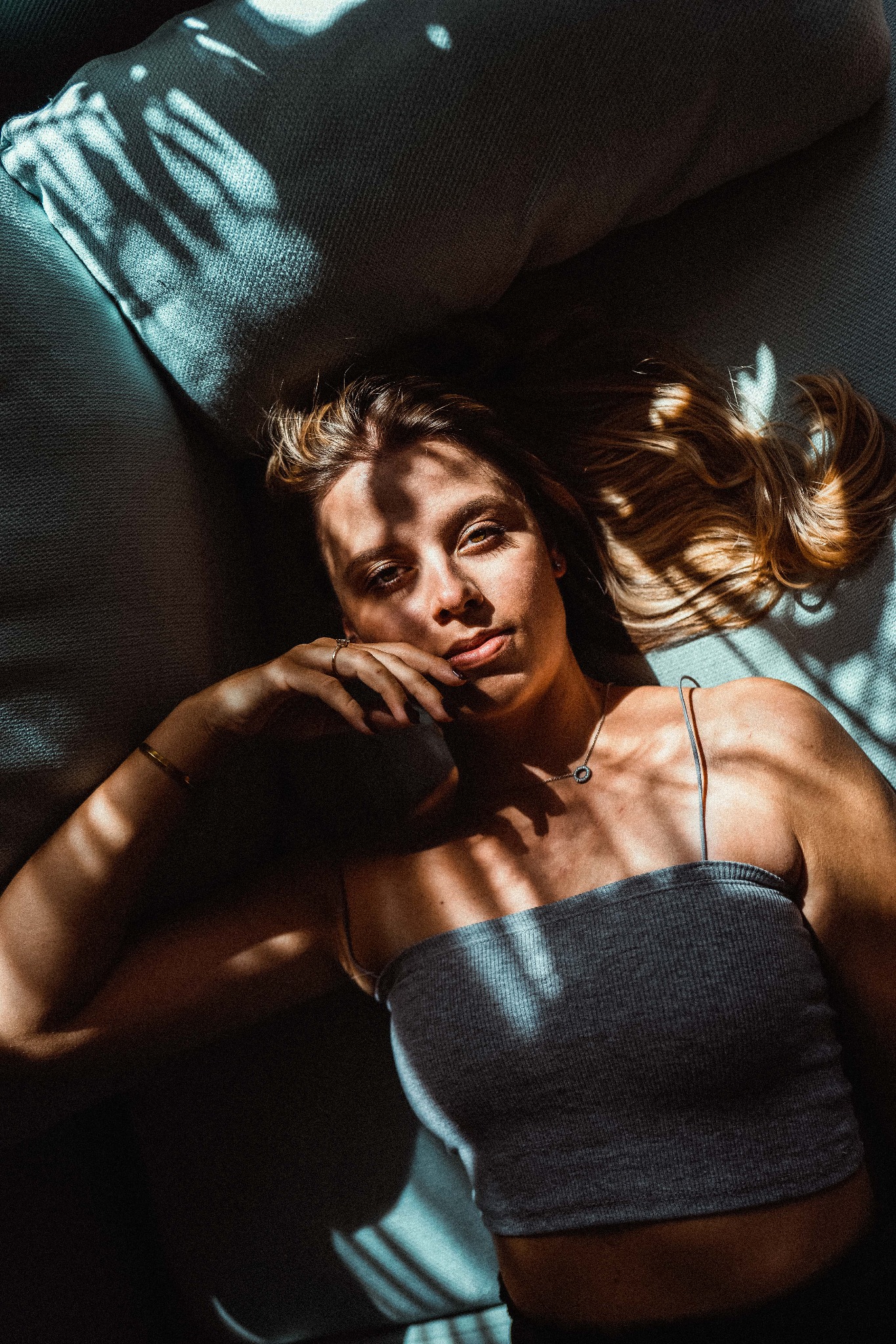 Portret kobiety leżącej na turkusowej kanapie w silnym, kontrastowym oświetleniu z cieniami liści na twarzy i ciele, z delikatnym uśmiechem i pierścionkiem na palcu.
