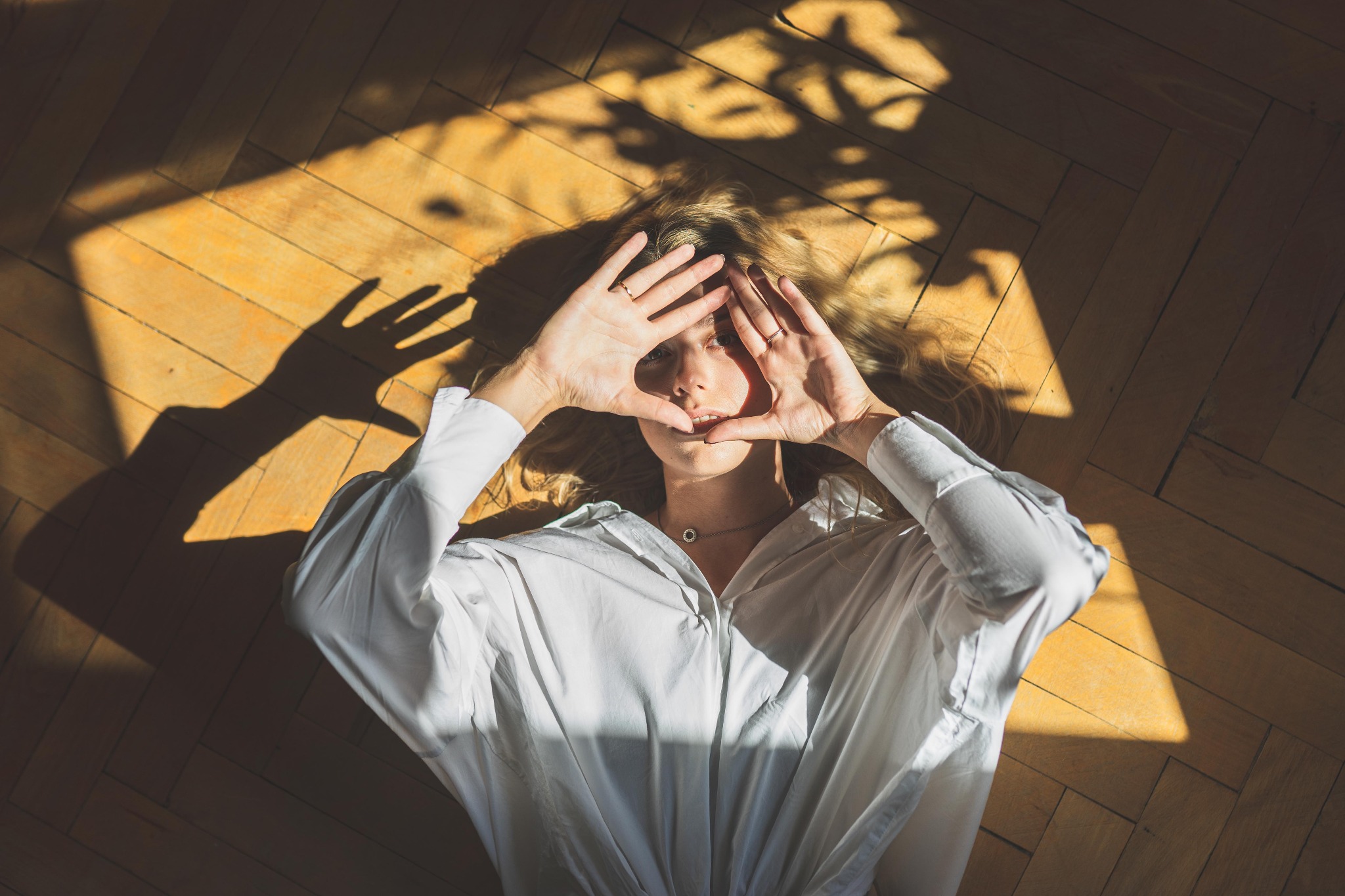 Kobieta w białej koszuli leży na parkiecie w promieniach słońca, dłońmi formuje ramkę wokół twarzy, widoczne pierścionki i naszyjnik