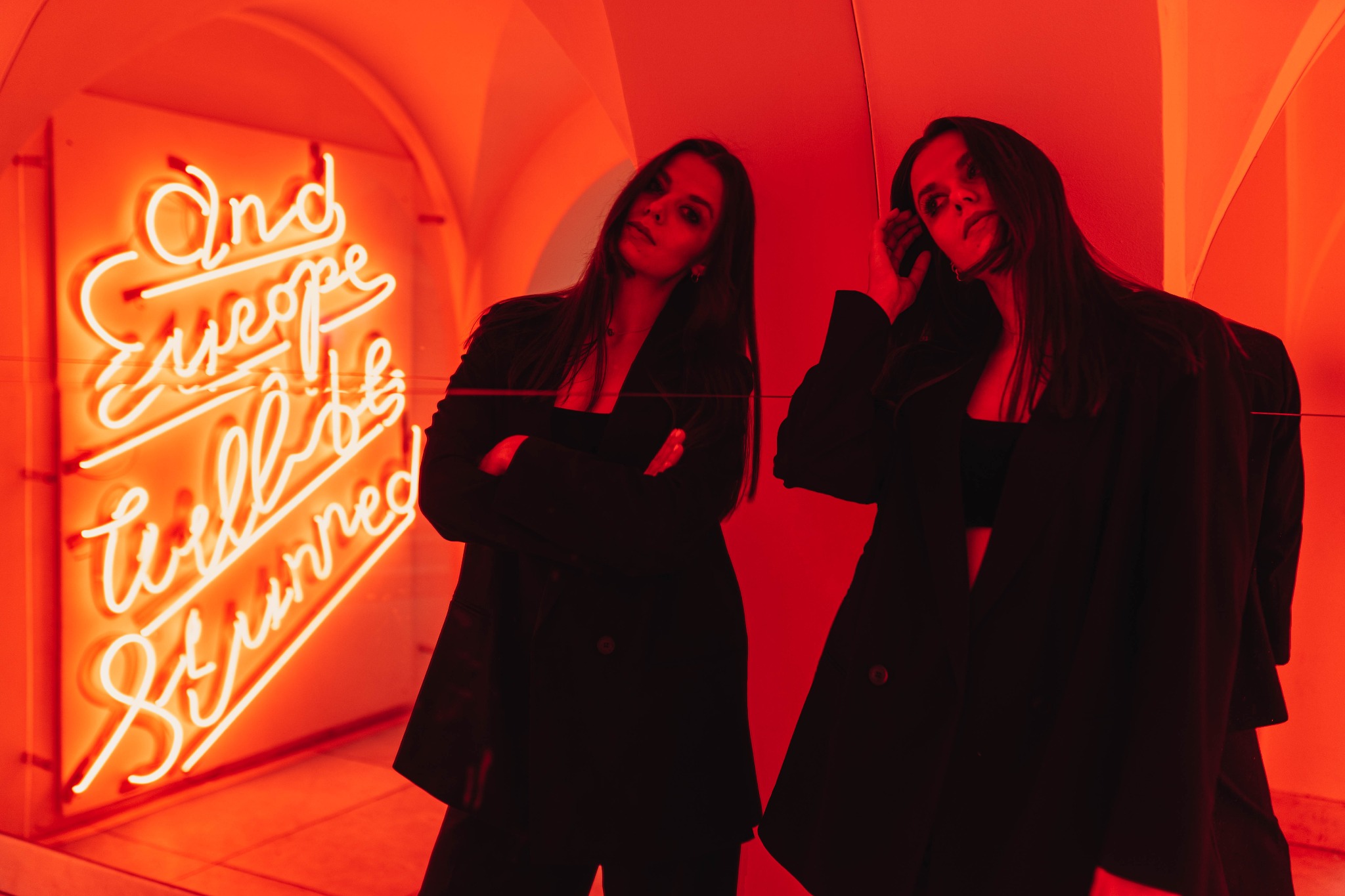 Kobieta w czarnym garniturze pozuje w czerwonym świetle obok neonu z napisem 'And Europe will be stunned', odbicie w lustrze.