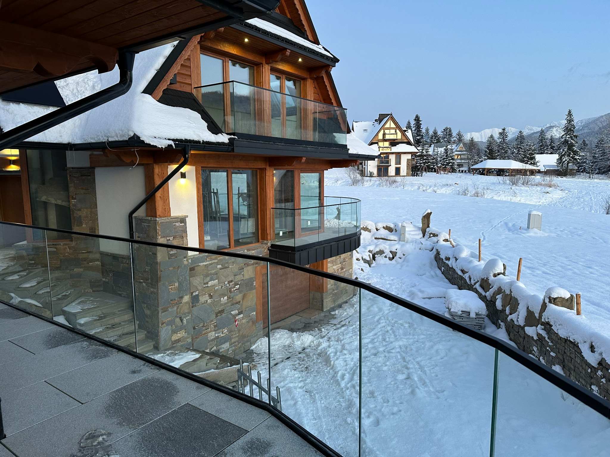 Nowoczesny dom w górskim stylu z przeszklonymi balkonami i tarasem, otoczony śnieżnym krajobrazem i kamiennym murkiem.