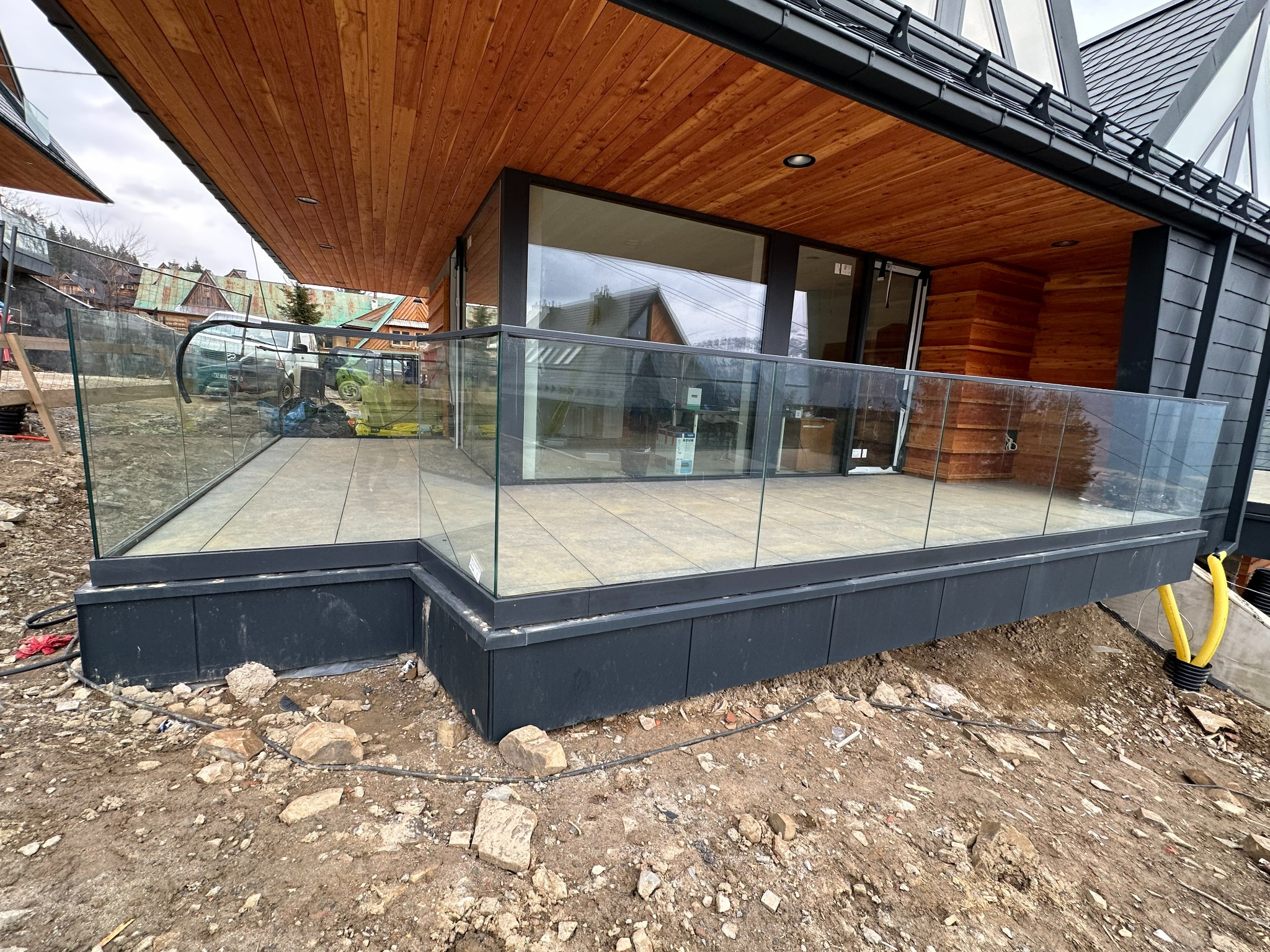 Nowoczesny balkon z szklaną balustradą w trakcie montażu, widoczne niezagospodarowane otoczenie i przewody instalacyjne.