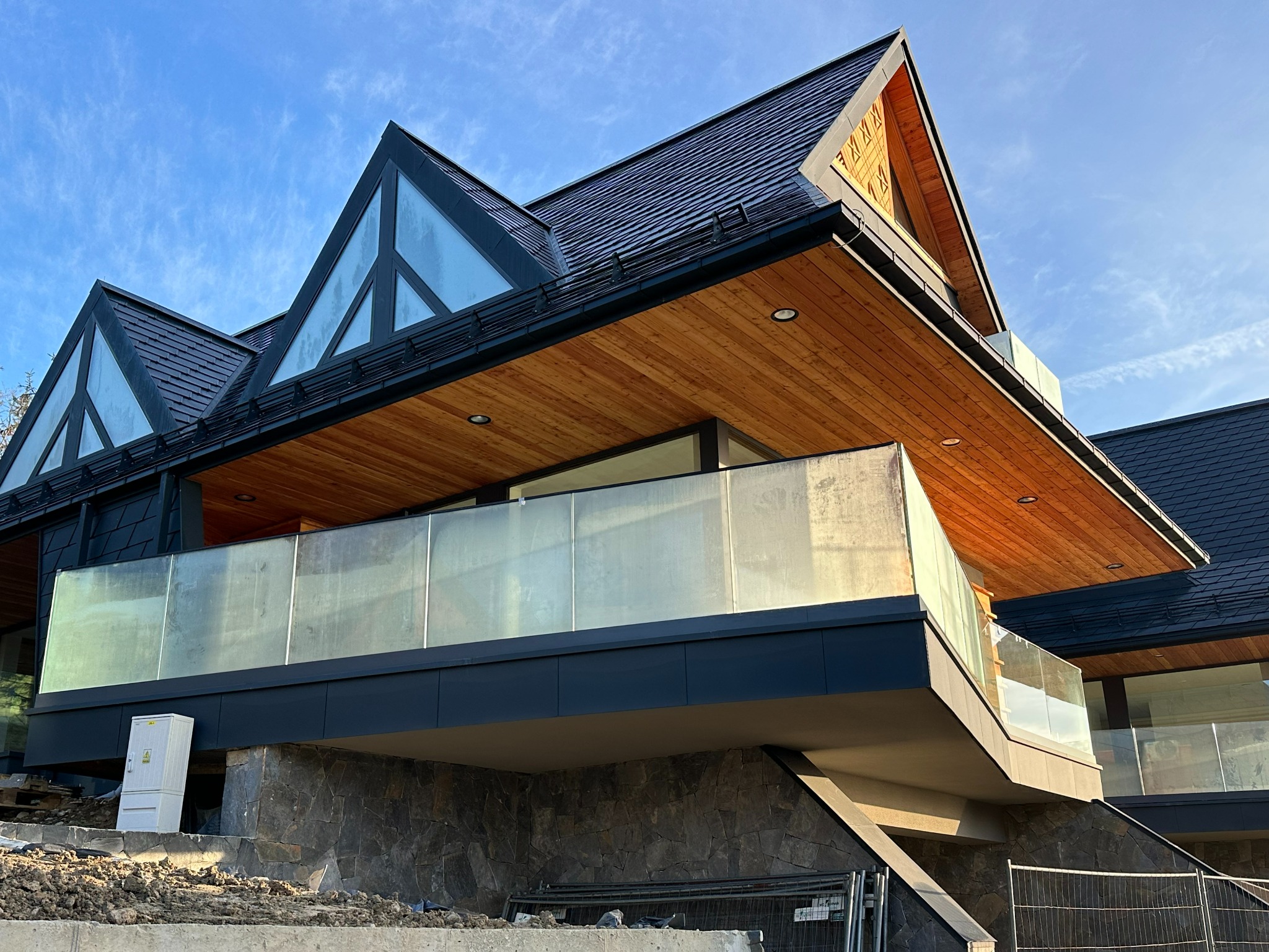 Nowoczesny dom z balkonem zabezpieczonym taflami szkła, widoczne drewniane wykończenia i elewacja z kamienia.