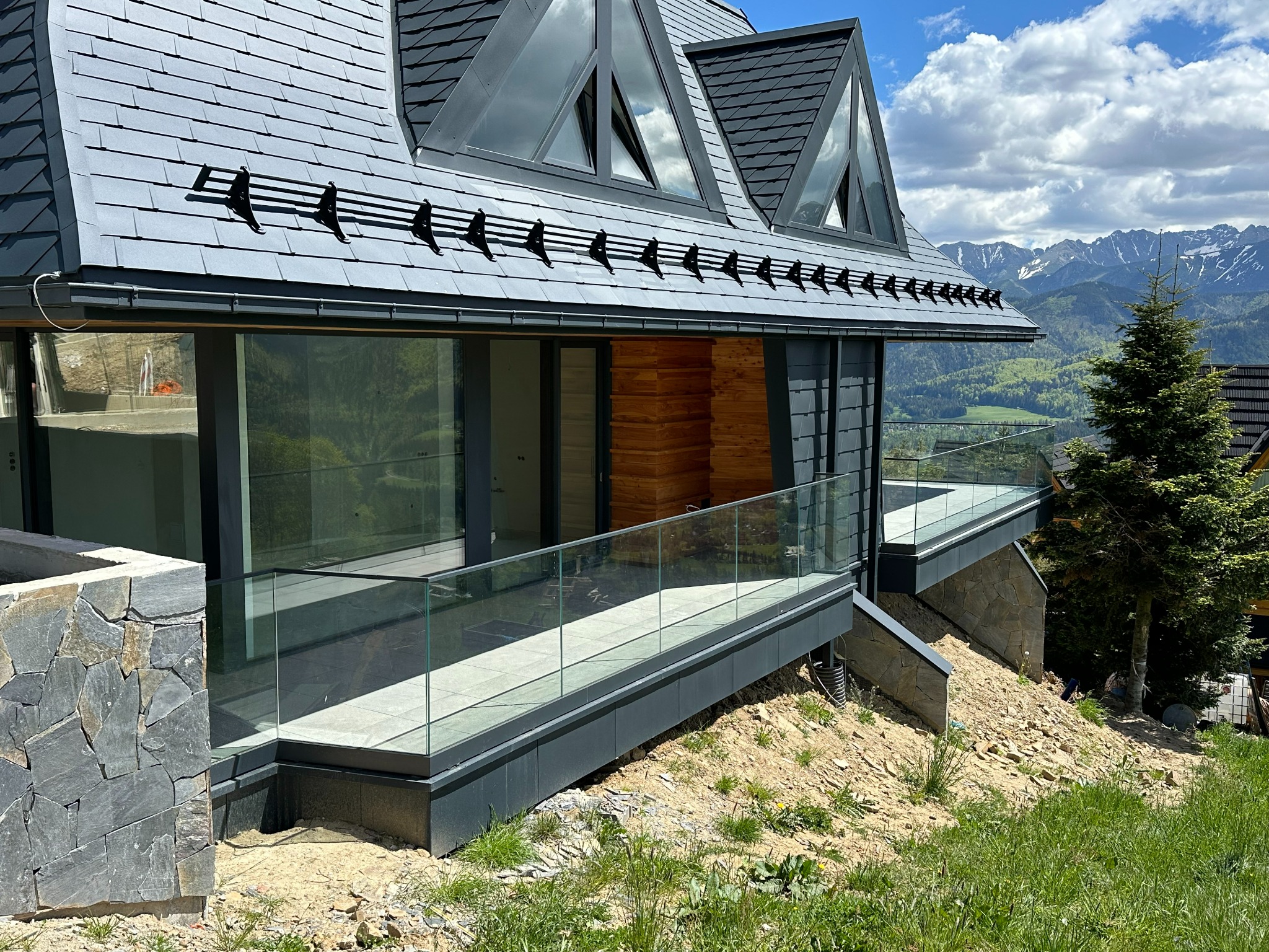 Nowoczesny dom z przeszklonym balkonem w Męcinie, z widokiem na góry i szklaną balustradą, otoczony zielenią.