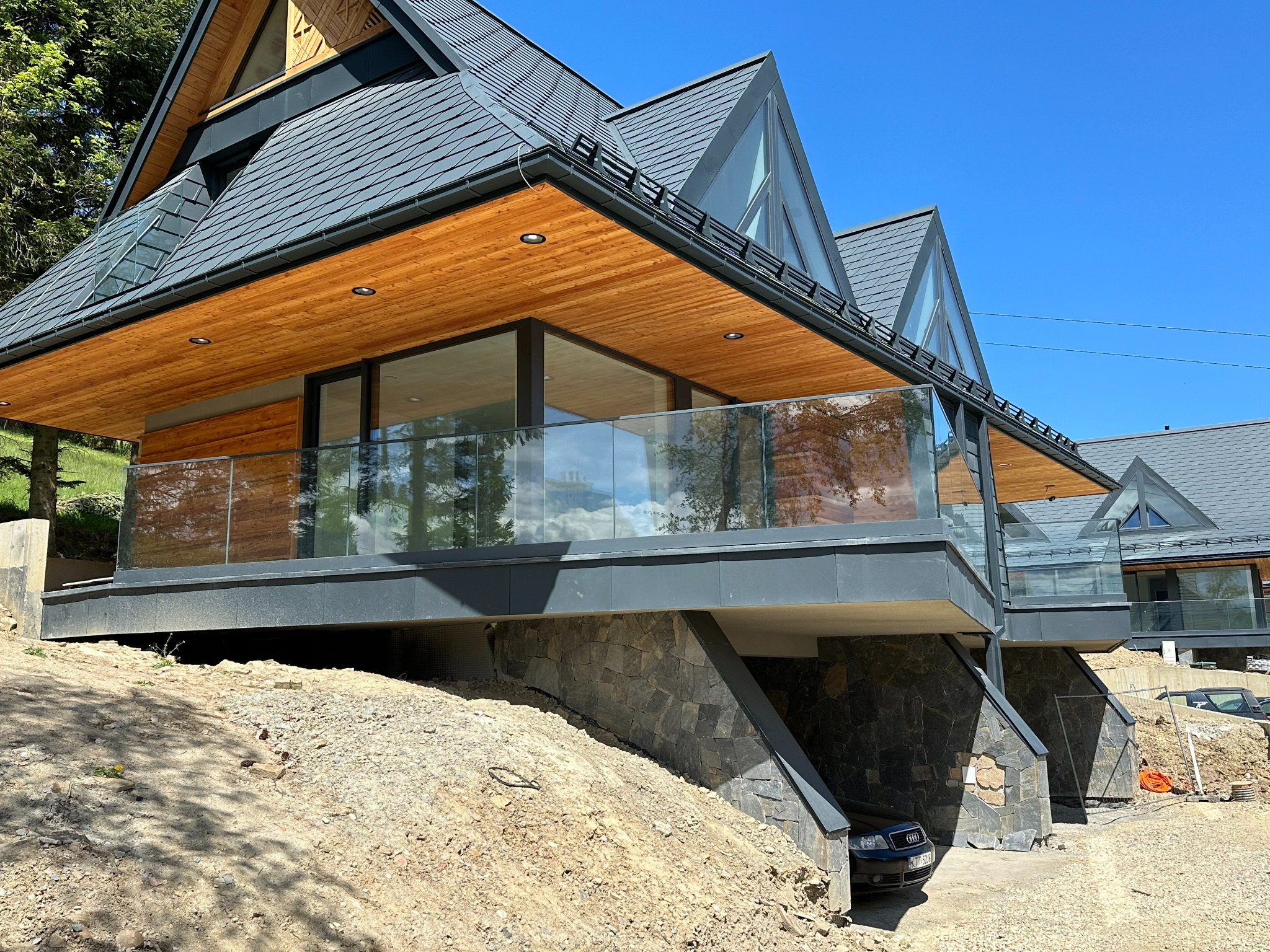 Nowoczesny dom w stylu góralskim z przeszklonymi balkonami i kamienną podmurówką, widok z dołu na tle błękitnego nieba.