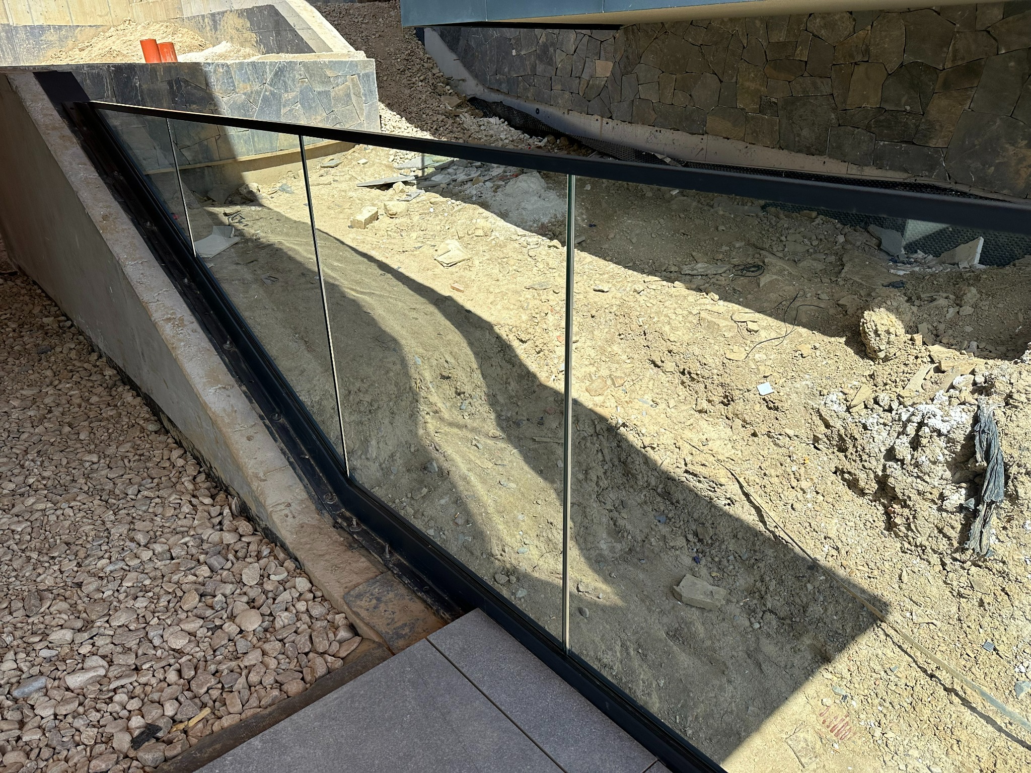 Szklana balustrada z czarną ramą, zamontowana na zewnątrz, widok na nierówny teren z ziemią i gruzem.