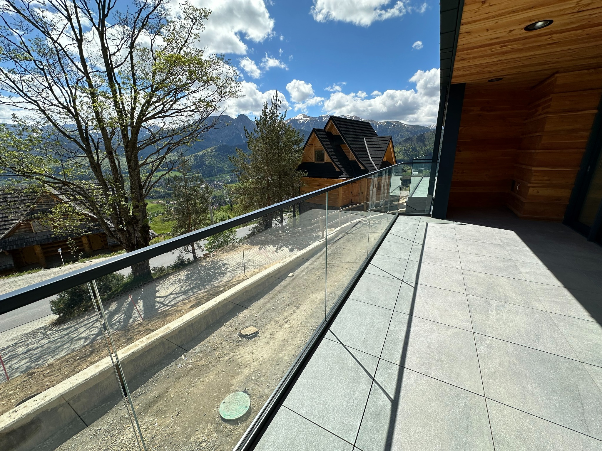 Szklana balustrada z czarnymi elementami mocującymi na tarasie z szarych płytek, widok na okoliczne domy i góry w tle.