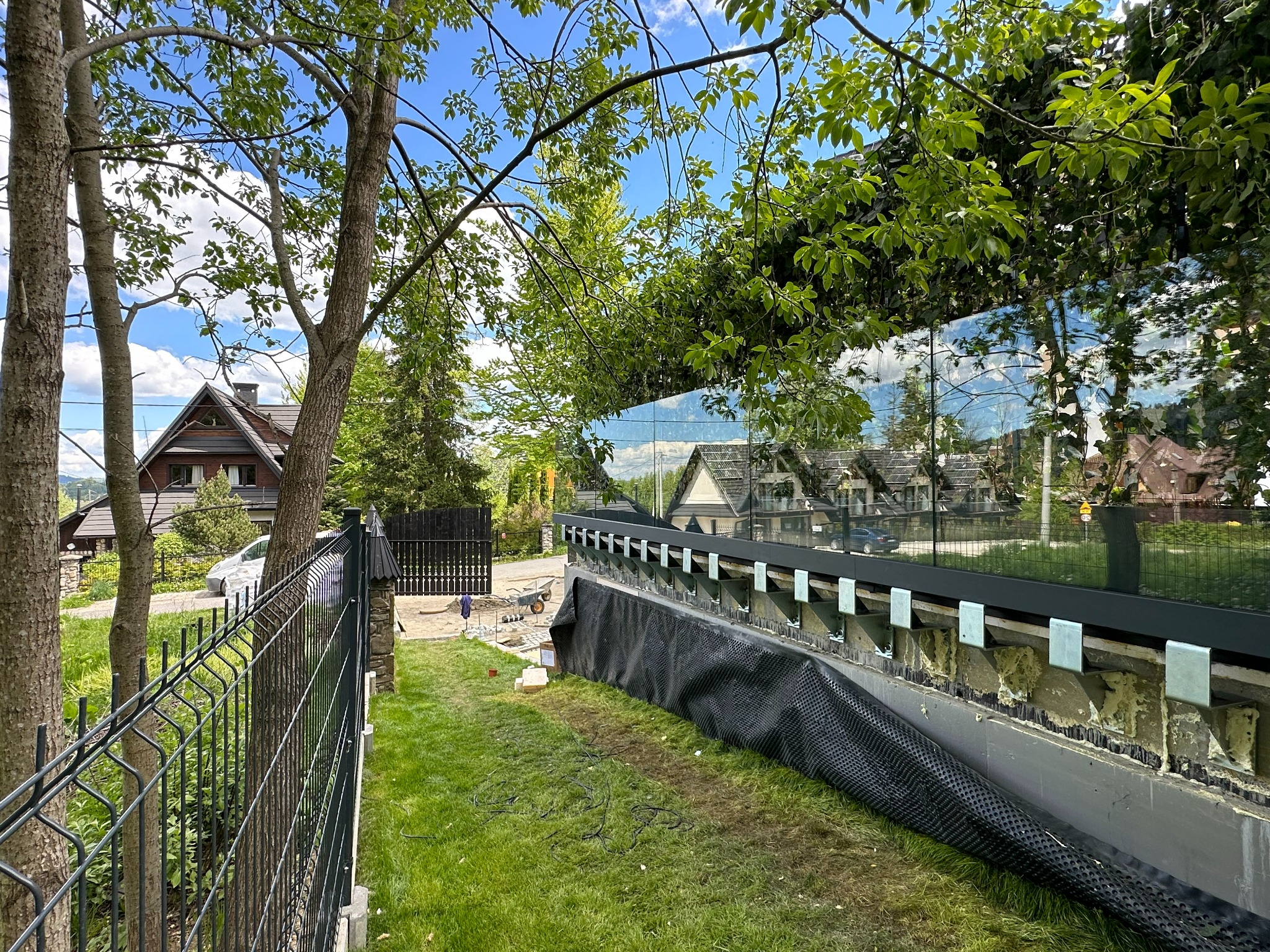 Widok z poziomu trawnika na montaż szklanej balustrady z czarnymi profilami, widoczne kotwy mocujące, czarna folia drenażowa, w tle dom i ogrodzenie panelowe.