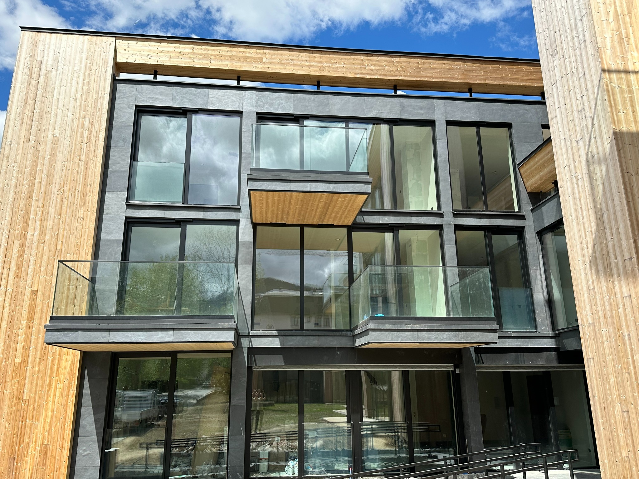 Nowoczesny budynek wielorodzinny z balkonami zabezpieczonymi szkłem, elewacja z szarego kamienia i drewnianymi panelami, błękitne niebo w tle.