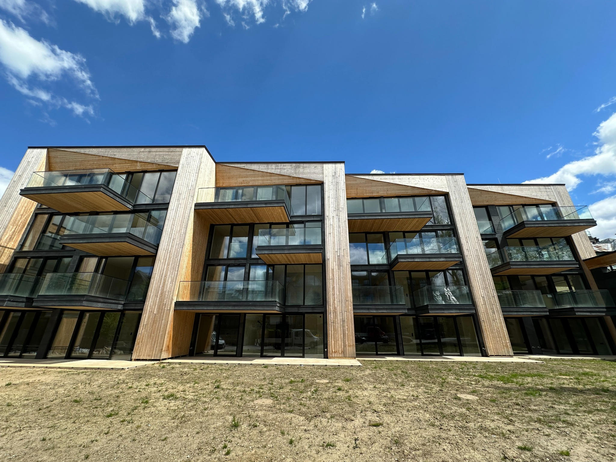 Nowoczesny budynek apartamentowy z drewnianą elewacją i balkonami z przezroczystymi taflami, widziany z dołu pod kątem, na tle błękitnego nieba z chmurami.