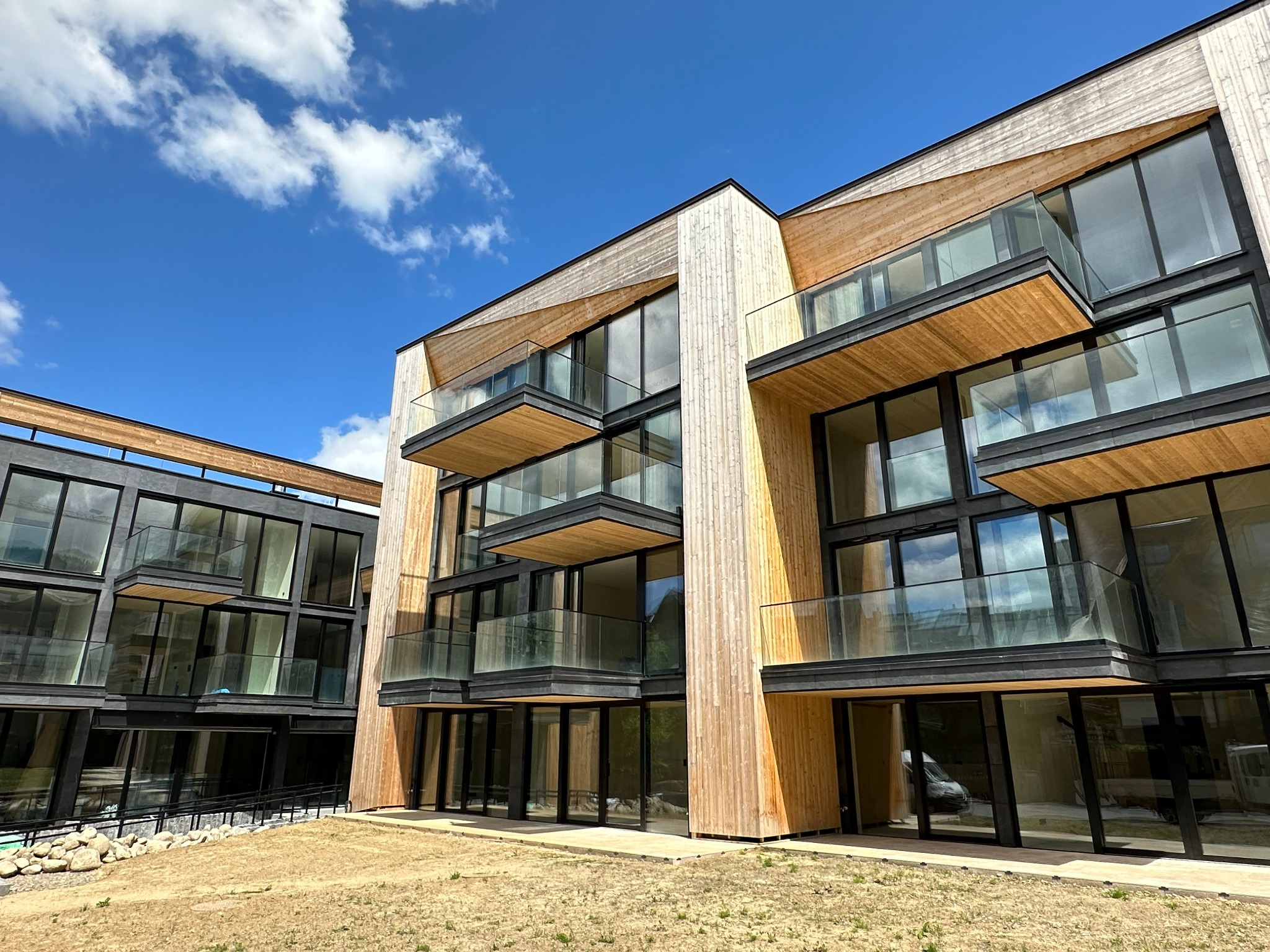 Nowoczesny budynek wielorodzinny z drewnianą elewacją i szklanymi balustradami balkonowymi na tle błękitnego nieba z chmurami, widok z dołu.