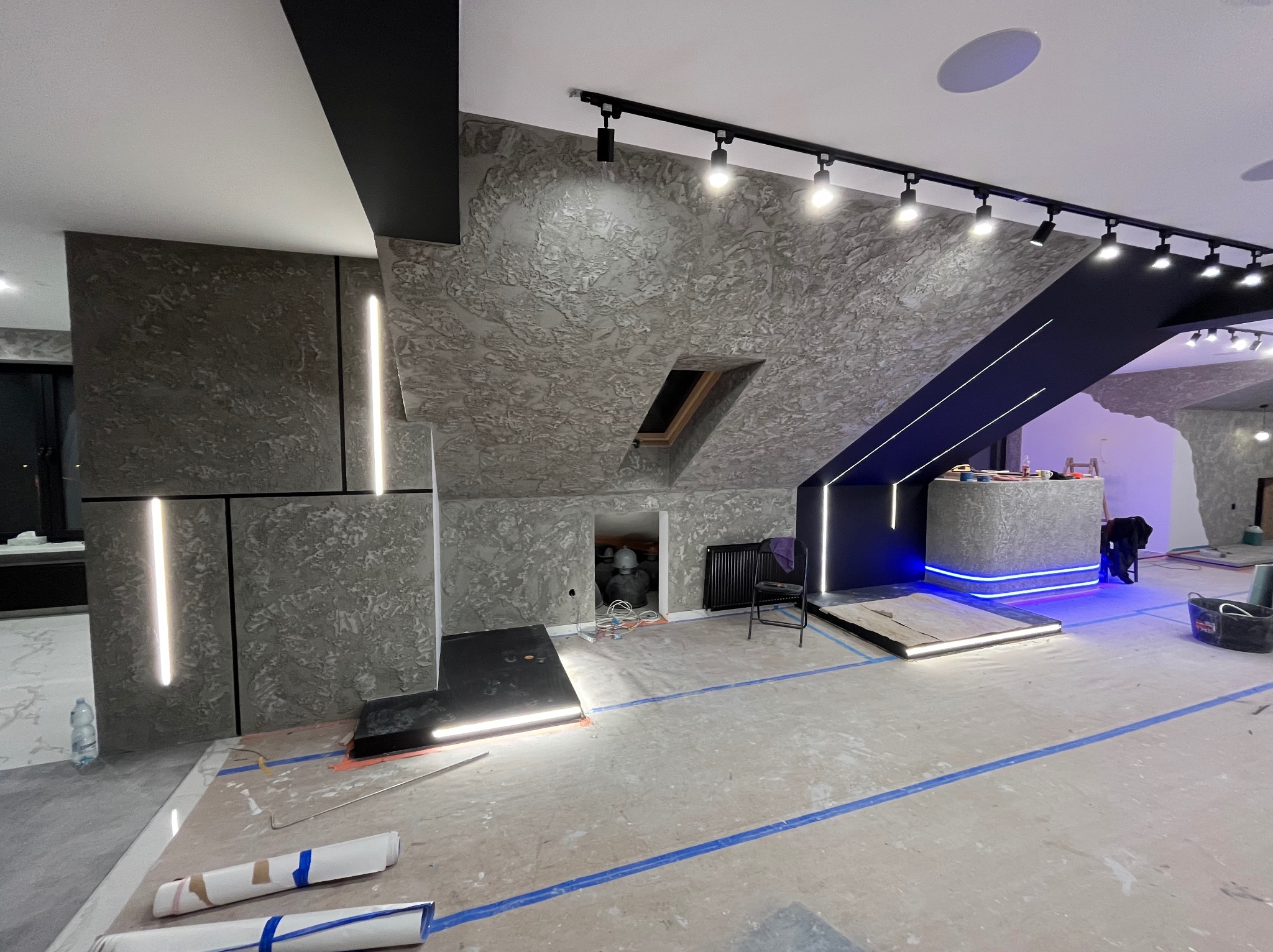 Wnętrze w trakcie remontu z surową, teksturowaną ścianą w odcieniach szarości, oświetleniem LED, przygotowaną podłogą i widocznym fragmentem zabudowy z podświetleniem.