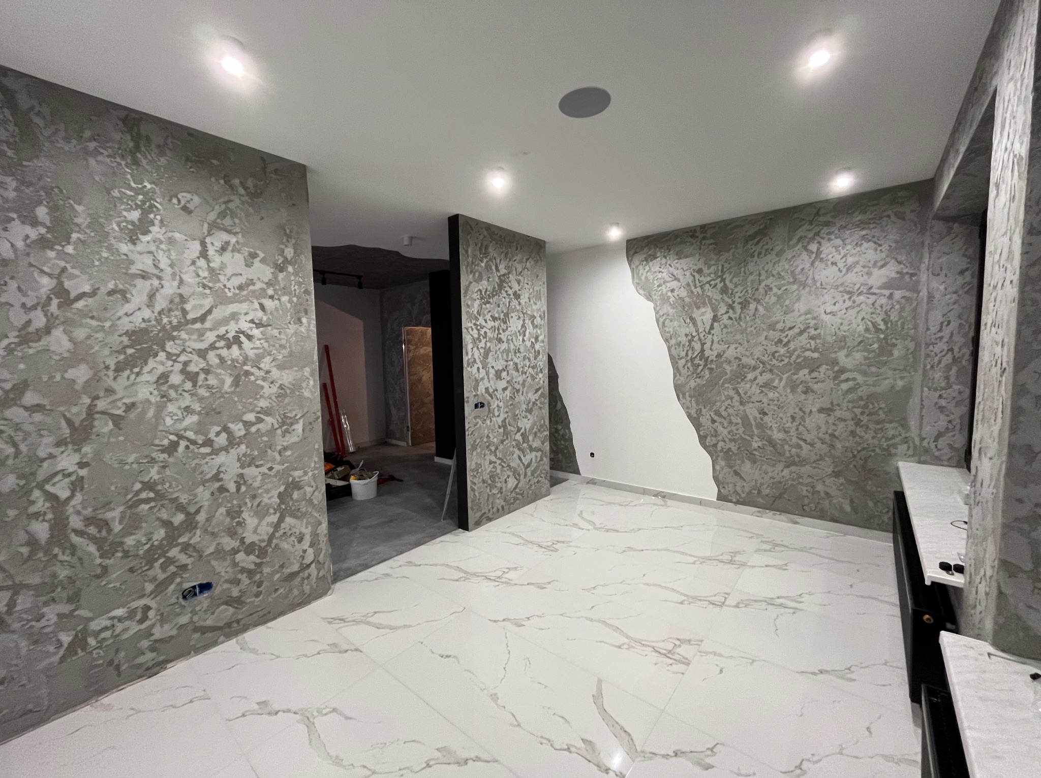 Wnętrze pokoju po remoncie: ściany pokryte dekoracyjną szpachlą w odcieniach szarości, biała marmurowa posadzka, widoczne narzędzia i wiadro w przejściu do innego pomieszczenia.