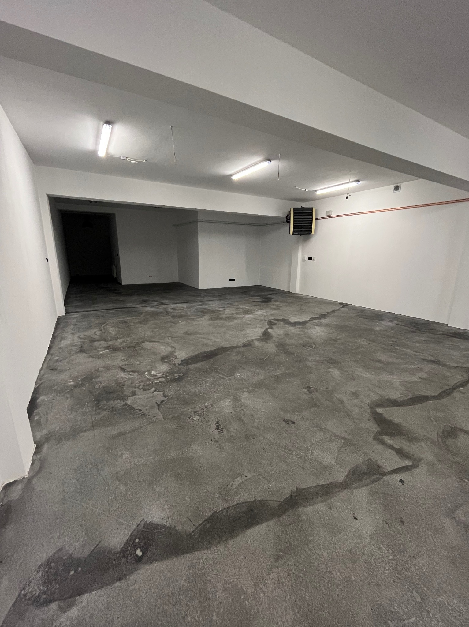 Wnętrze hali z surową, szarą posadzką żywiczną w trakcie schnięcia, widoczne ślady wilgoci i refleksy światła, białe ściany i sufit z lampami, wentylator pod sufitem.