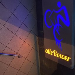 Bestprom Producent Reklam - Neonowy szyld z niebieską sylwetką rowerzysty i żółtym napisem 'alleRower' na ścianie z ukośnymi płytkami, oświetlony niebieskim światłem.