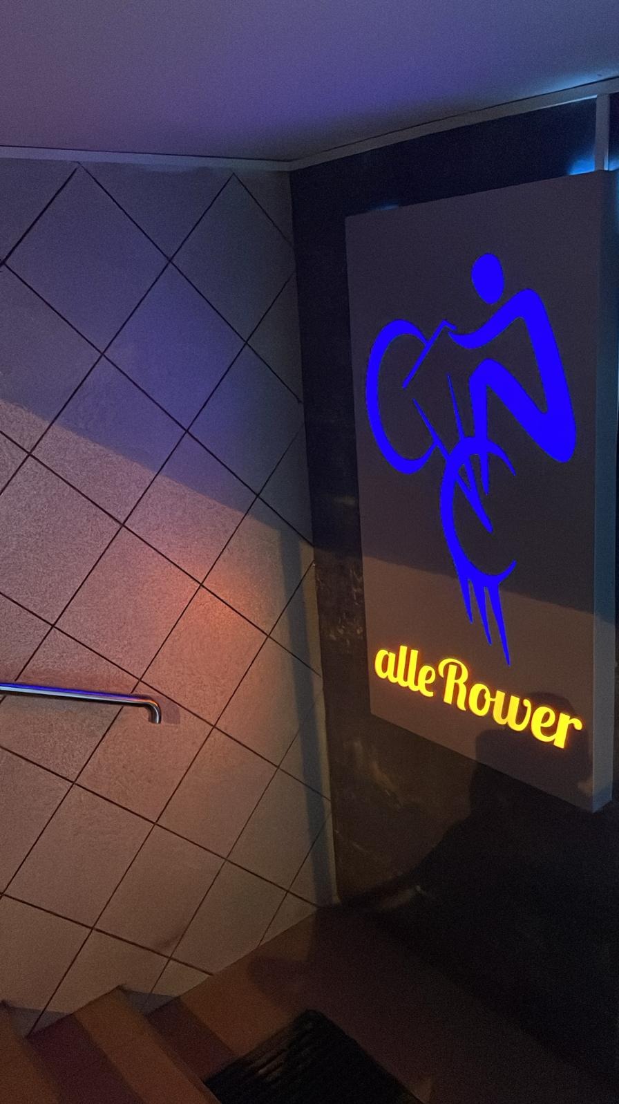 Neonowy szyld z niebieską sylwetką rowerzysty i żółtym napisem 'alleRower' na ścianie z ukośnymi płytkami, oświetlony niebieskim światłem.