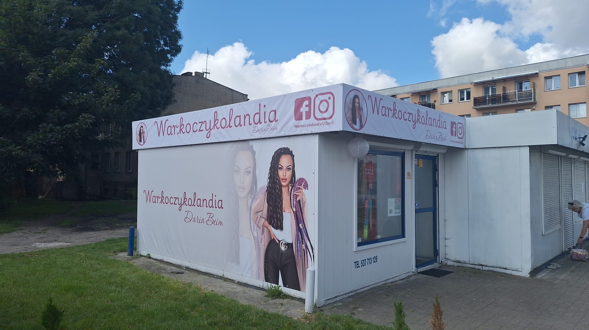 Oklejony pawilon z reklamą salonu fryzjerskiego 'Warkoczykolandia' z wizerunkiem kobiety z warkoczami i danymi kontaktowymi. Widoczne logo i ikony mediów społecznościowych.