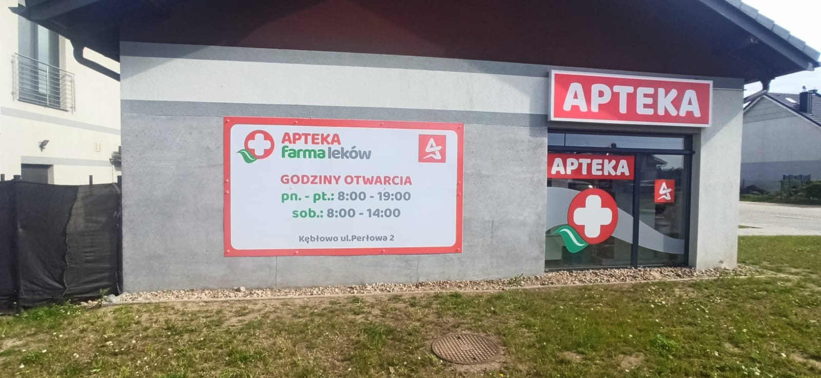 Oklejona witryna apteki z logo i godzinami otwarcia. Na ścianie budynku duży baner informacyjny z nazwą i danymi kontaktowymi. Widoczne elementy zieleni.
