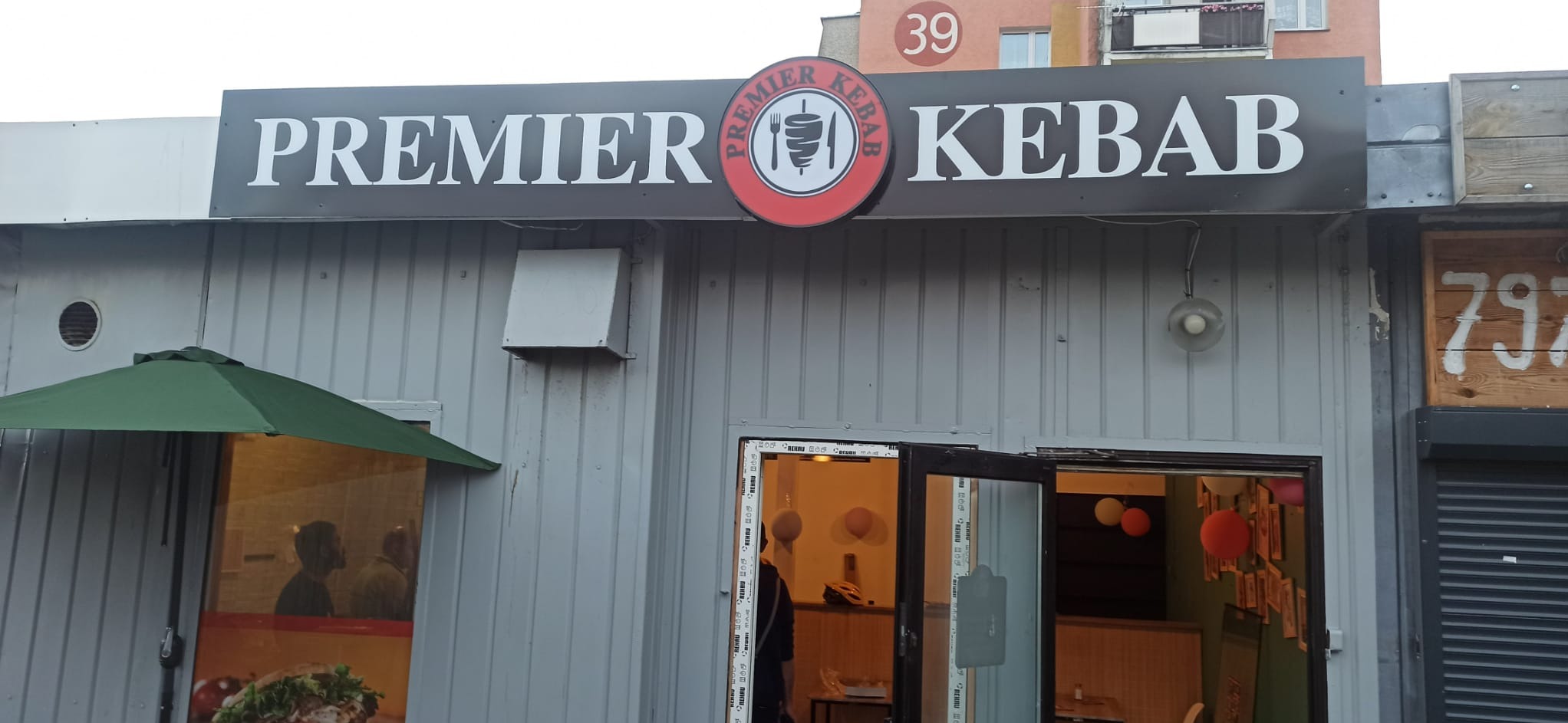 Szary budynek z szyldem 'Premier Kebab' z logo z kebabem na tle numeru budynku 39. Widoczne otwarte drzwi i okno z zielonym parasolem.