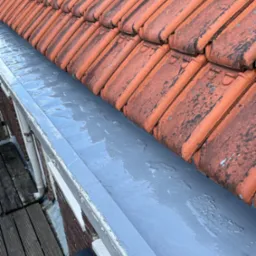 Świeżo pomalowana, szara rynna pod brudnymi, czerwonymi dachówkami. Widoczne ślady pędzla i niedoskonałości na powierzchni rynny. W tle fragment drewnianego tarasu.