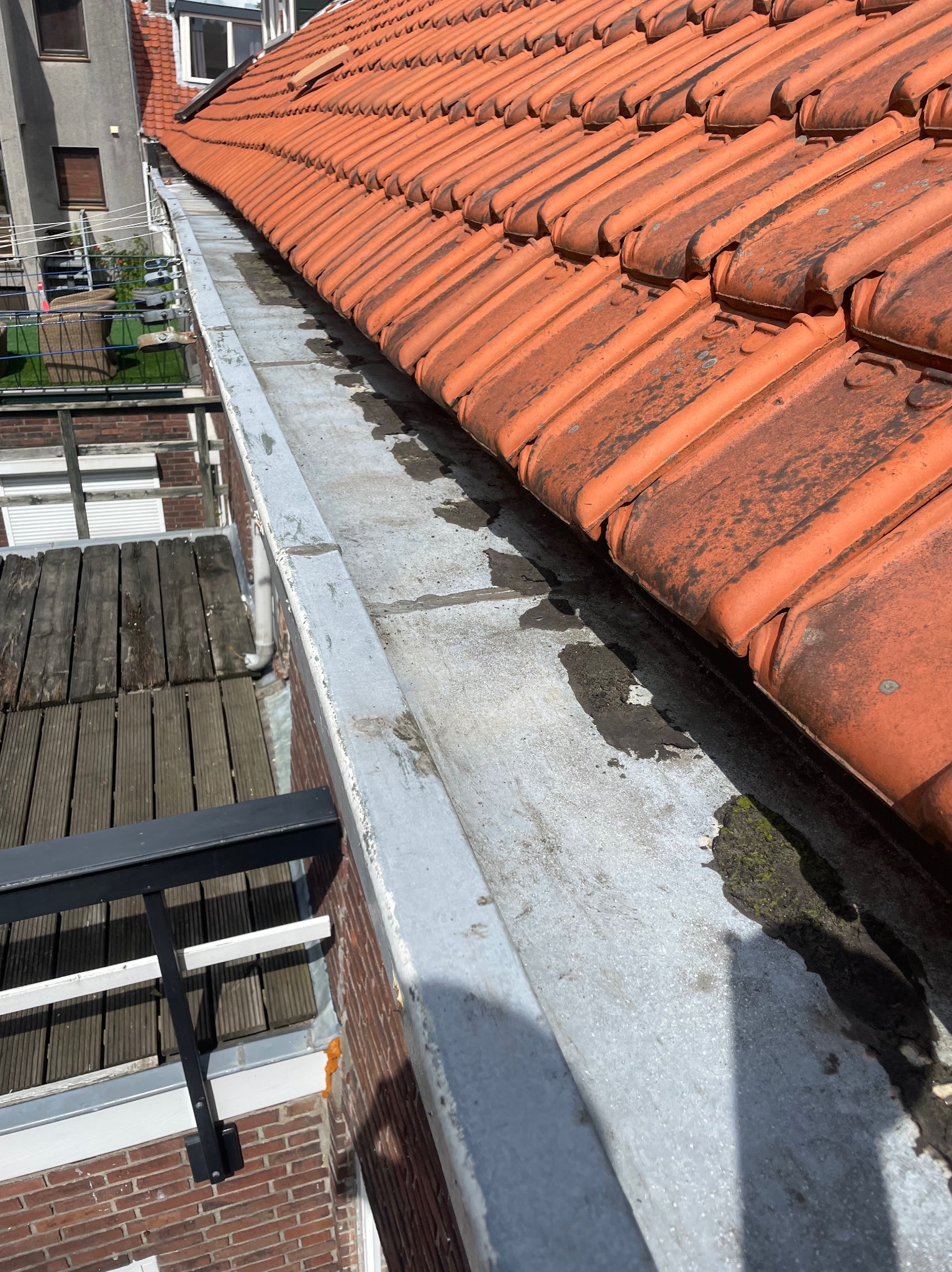 Częściowo oczyszczona, szara rynna z widocznymi śladami wilgoci i mchu, przylegająca do pomarańczowego dachu z dachówki ceramicznej, z widocznym fragmentem ceglanego budynku i drewnianego balkonu...