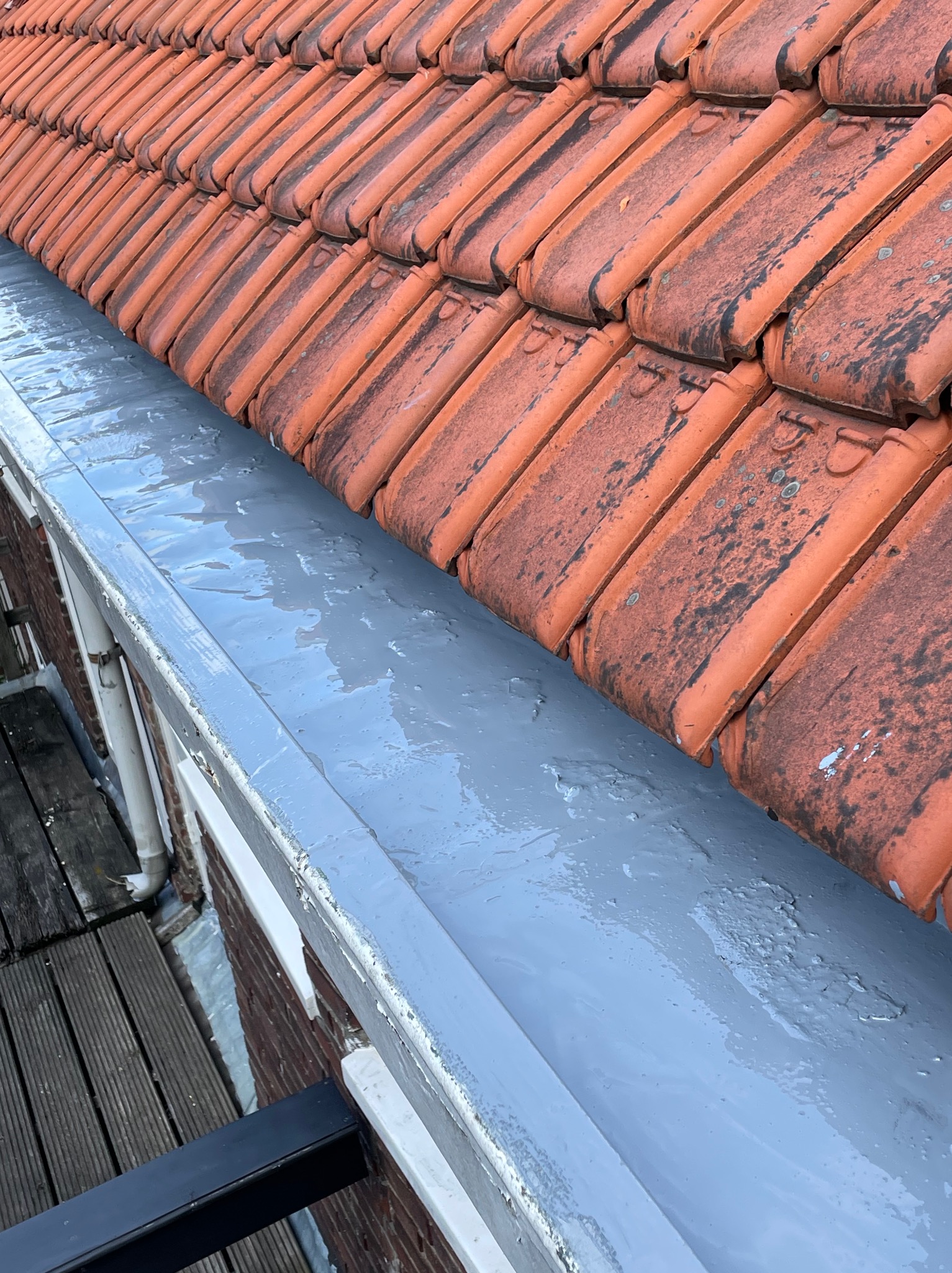 Świeżo pomalowana, szara rynna pod brudnymi, czerwonymi dachówkami. Widoczne ślady pędzla i niedoskonałości na powierzchni rynny. W tle fragment drewnianego tarasu.