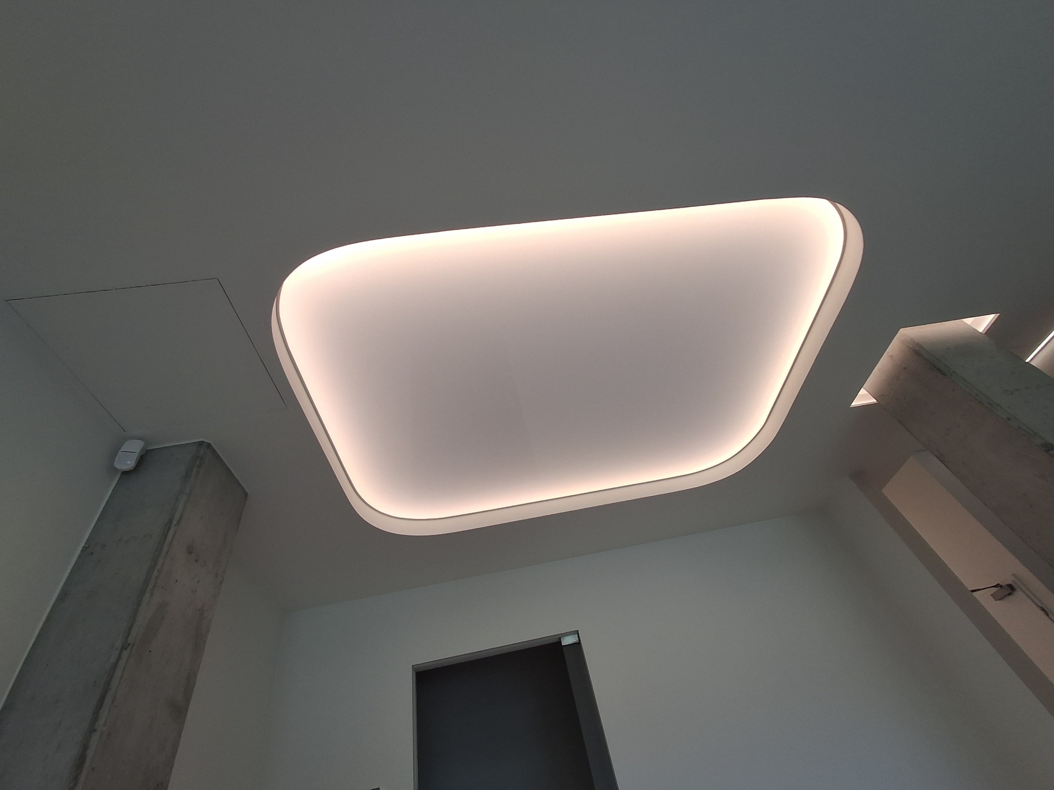 Nowoczesny sufit z podświetleniem LED w kształcie zaokrąglonego prostokąta, widok z dołu, z fragmentem betonowego filaru i drzwiami w tle. Minimalistyczny design.